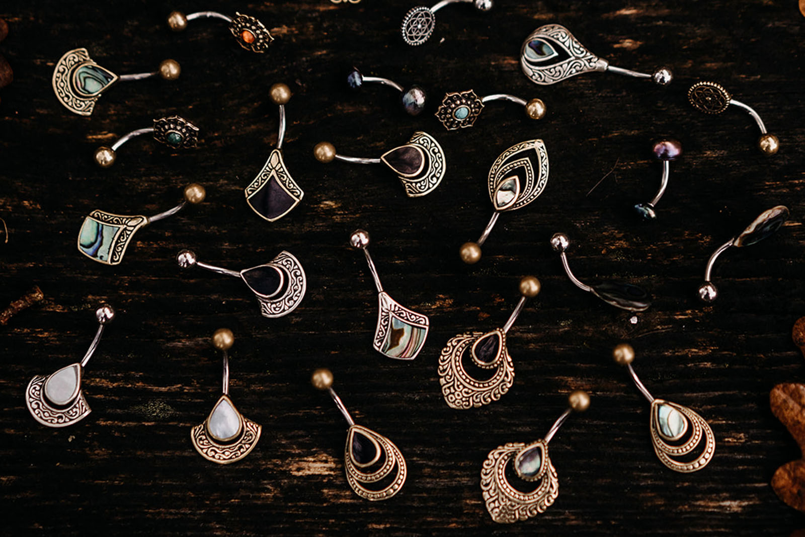 Viele goldene und silberne Bauchnabelpiercings mit orientalischen hippie Muster liegen auf einer dunkelbraunen Holzbank umgeben von Eichenblaettern