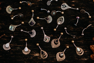 Viele goldene und silberne Bauchnabelpiercings mit orientalischen hippie Muster liegen auf einer dunkelbraunen Holzbank umgeben von Eichenblaettern