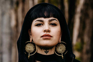 Weisse dunkelhaarige Frau mit Pony und tattoowiertem Hals schaut selbstsicher in die Kamera und trägt eine schwarze Kapuze und goldene grosse Mandala Ohrringe in gedehnten Ohren während sie vor einer Holzwand steht